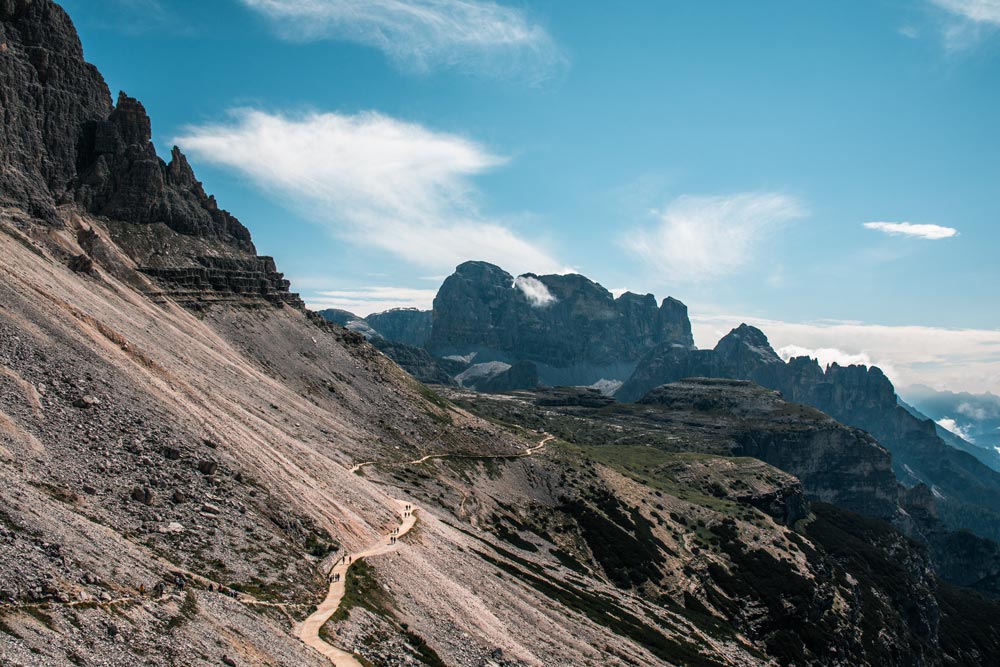 Blick auf den Start der drei Zinnen Wanderung oberhalb der Auronzohütte. Der Wanderweg schlängelt sich an dem Berg entlang. In der Ferne ist ein weitläufiges Gebirge zu sehen.